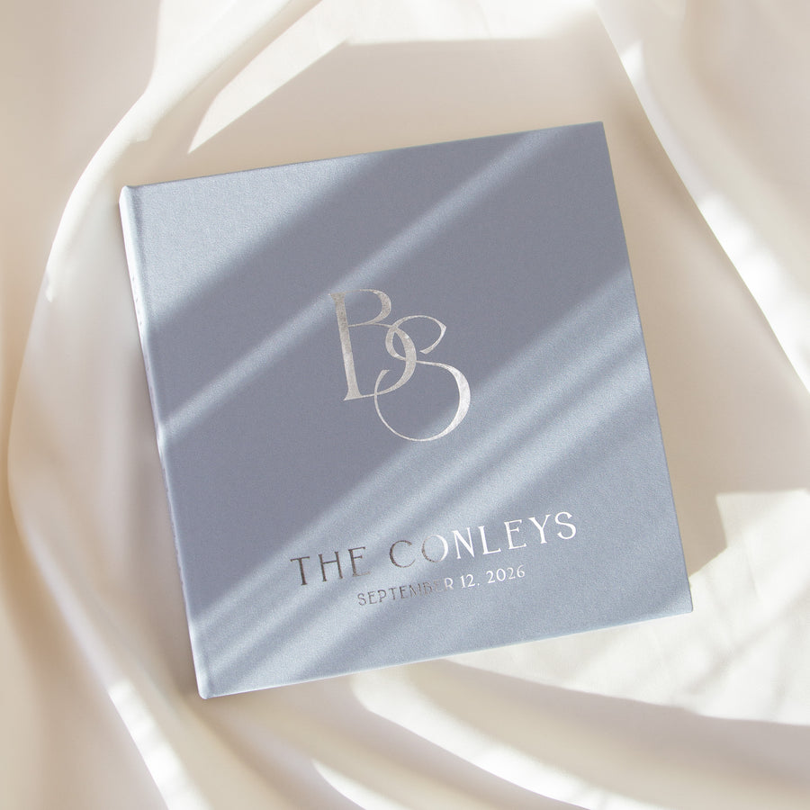 A square light blue keepsake album or photo book with a metallic silver monogram logo and the text 'THE CONLEYS' and 'SEPTEMBER 9, 2026,' styled on draped cream satin fabric with soft diagonal shadows.