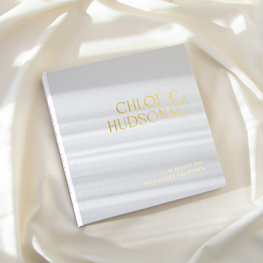 Silky White Polaroid wedding guest book with Real Gold personalization styled on flowing ivory fabric in natural light.