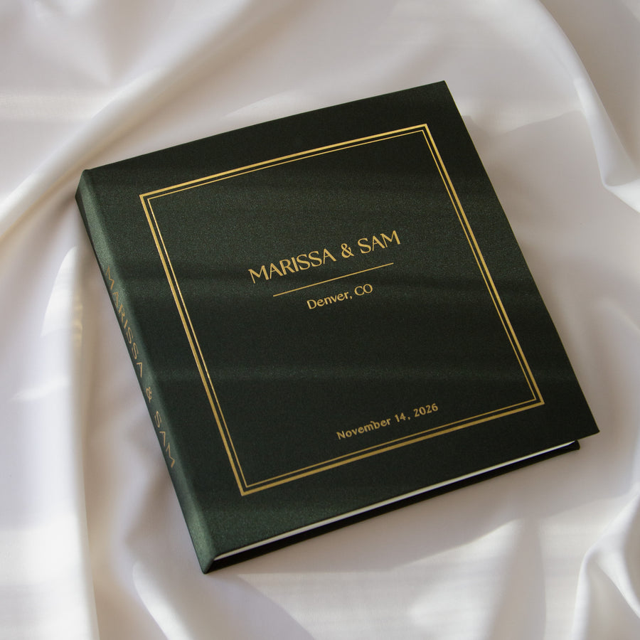 A personalized hardcover photo album in dark forest green with elegant gold foil lettering and border details, resting on draped white satin fabric.
